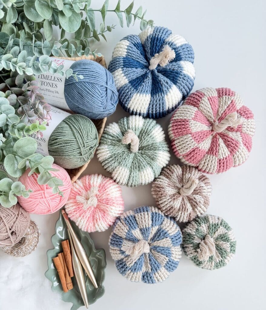 Several crocheted pumpkins in various colors, including checkerboard and striped patterns, are arranged next to yarn balls, cinnamon sticks, and decorative greenery.