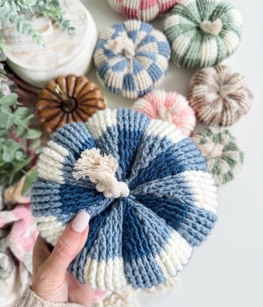 Hand holding a blue and white striped crochet pumpkin, surrounded by other small knit and wooden decorative pumpkins.