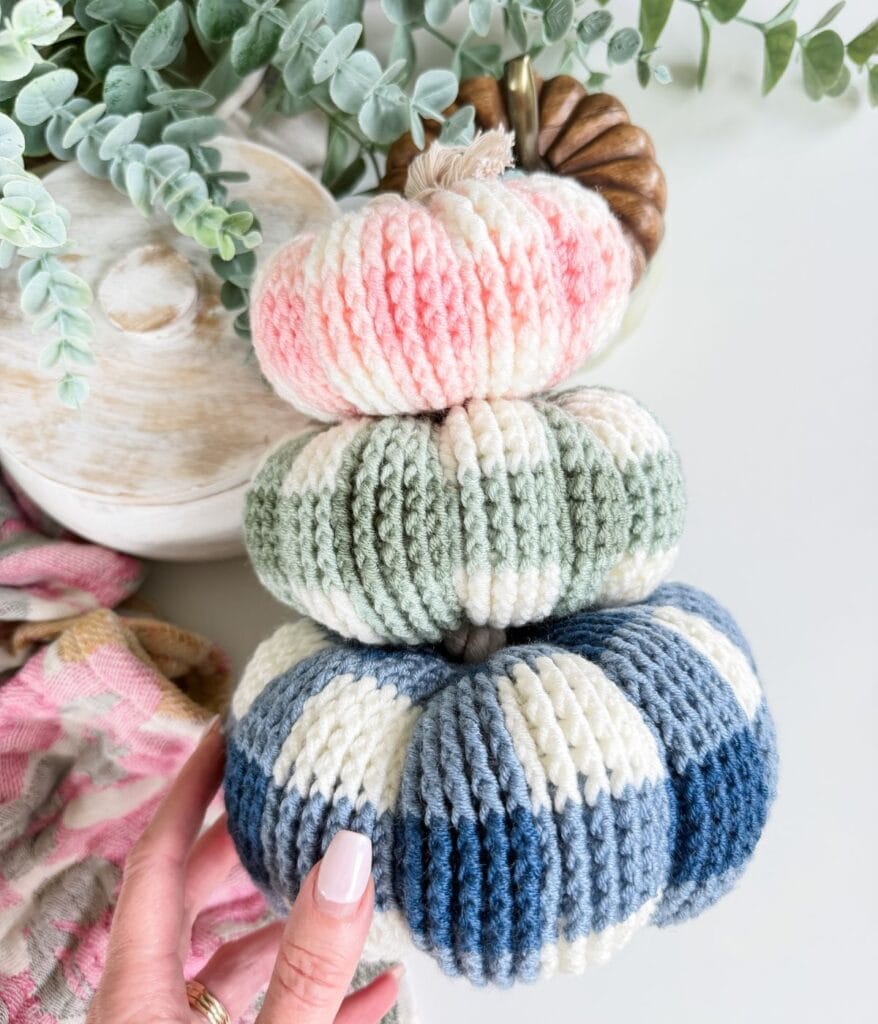 A hand holding a stack of three small crochet pumpkins in shades of pink, green, and blue, each featuring a classic checker pumpkin pattern.