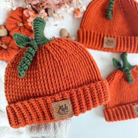 Three orange crochet pumpkin hats, each topped with a green stem and leaf, and featuring a brown leather brand tag.