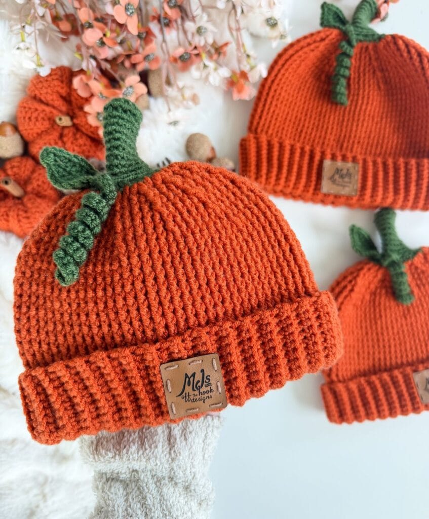 Three orange crochet pumpkin hats, each topped with a green stem and leaf, and featuring a brown leather brand tag.