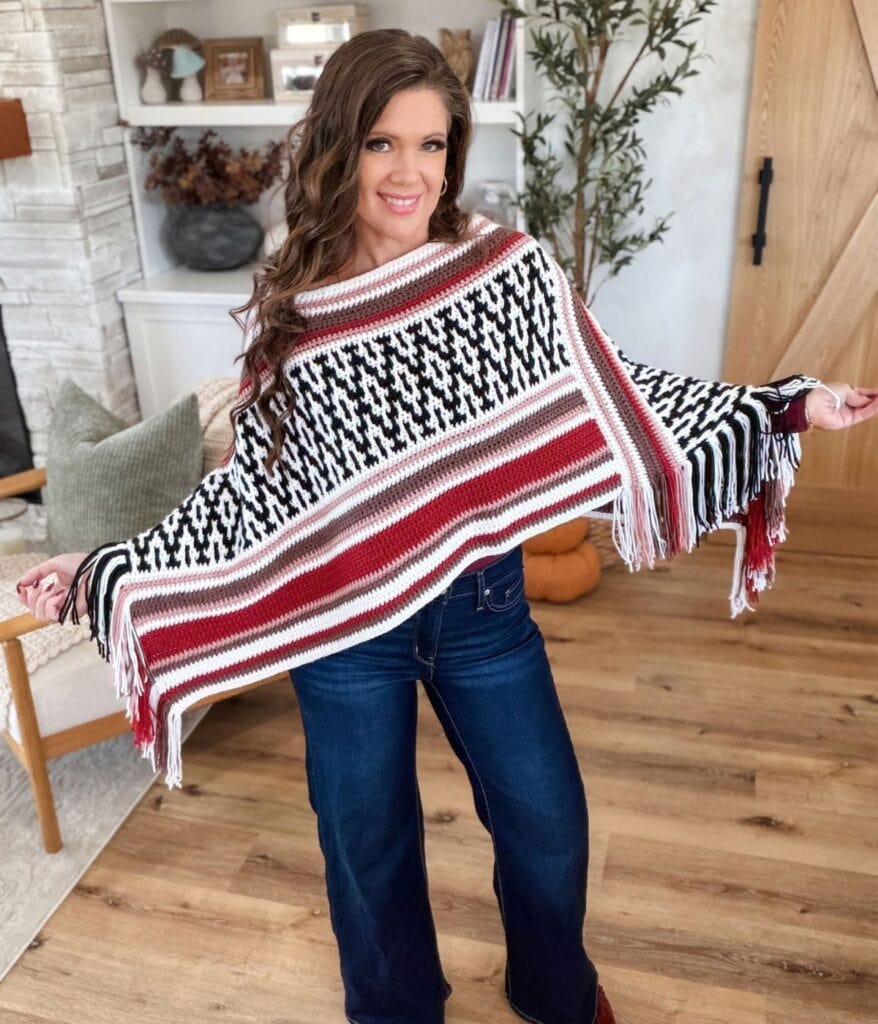 A woman wearing a black, white, and maroon mosaic crochet poncho pattern with fringe, paired with blue jeans.