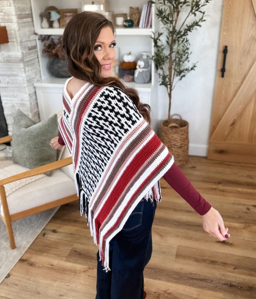Viewed from behind, a woman wears a black, white, and burgundy mosaic crochet poncho pattern with fringe.