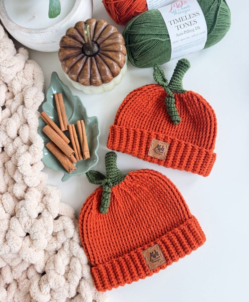 Two orange, hand-knitted pumpkin hats with green stems and branded tags, displayed with a small ceramic pumpkin, cinnamon sticks, and a skein of green yarn for a crochet pumpkin hat project.