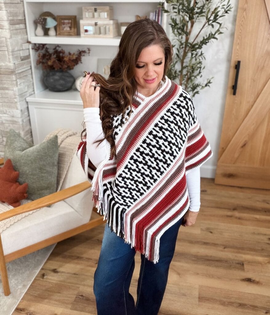 A woman wears a white long-sleeve shirt under a black, white, and maroon mosaic crochet poncho pattern with fringe trim and blue jeans.