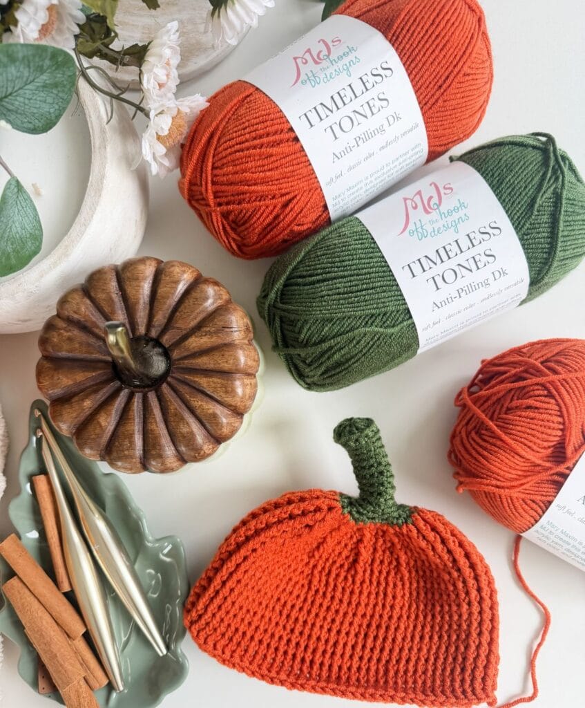 Two skeins of orange and green yarn, a crochet pumpkin, a wooden pumpkin decoration, and cinnamon sticks on a white surface, perfect for a pumpkin hat pattern.