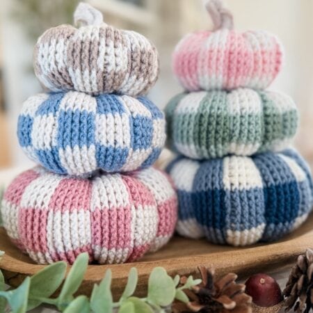 Two stacks of crocheted gingham pumpkins in pastel colors arranged on a wooden tray with pinecones and greenery.