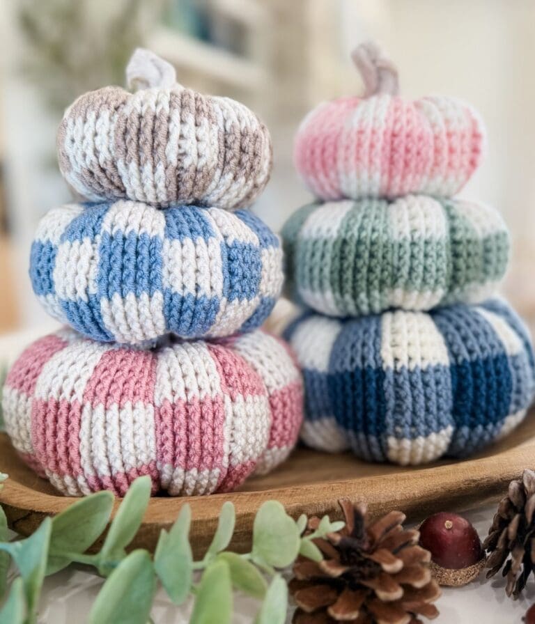 Two stacks of crocheted gingham pumpkins in pastel colors arranged on a wooden tray with pinecones and greenery.