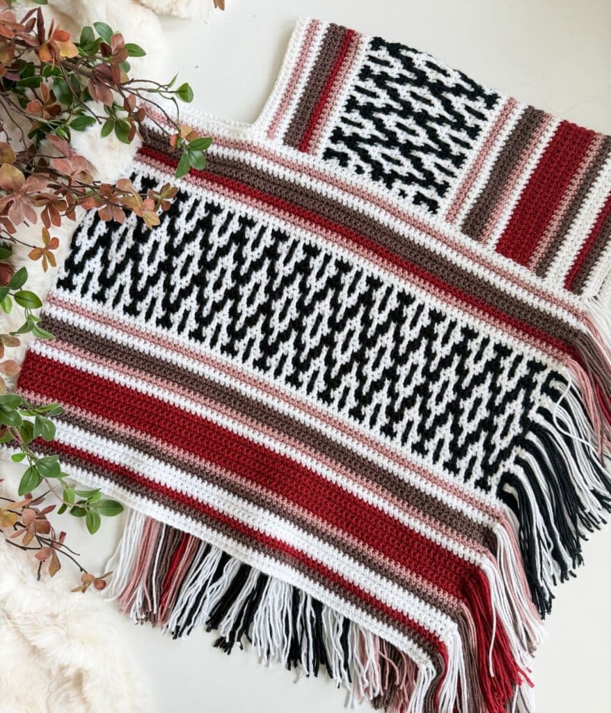 Crochet poncho in a geometric mosaic crochet pattern featuring black, white, red, brown, and pink stripes with fringe along the hem.