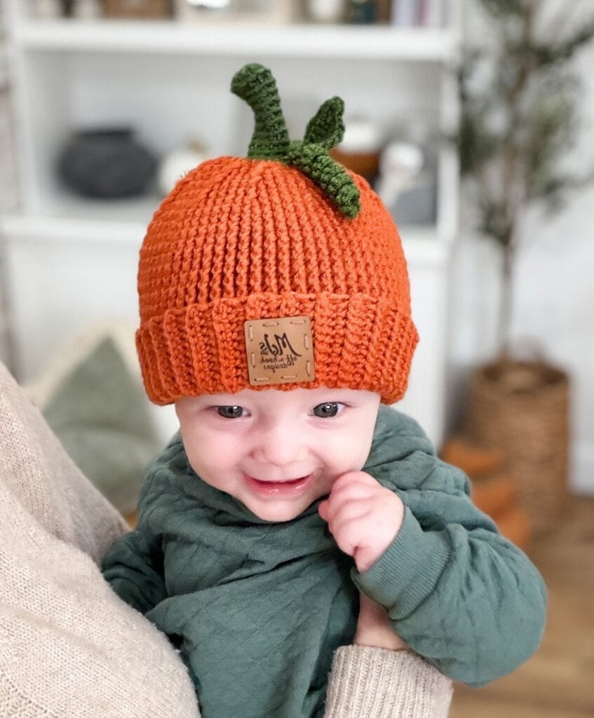 A baby wearing a crochet pumpkin hat with a green stem, featuring a detailed pumpkin hat pattern.