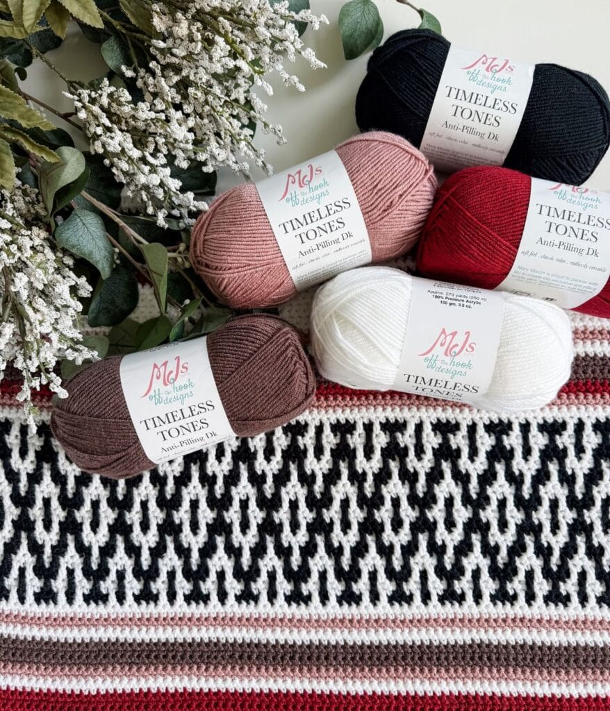 Five skeins of MJ’s Off the Hook Designs Timeless Tones yarn in black, pink, red, brown, and white, arranged for a mosaic crochet poncho pattern on a knit blanket with white floral sprigs.