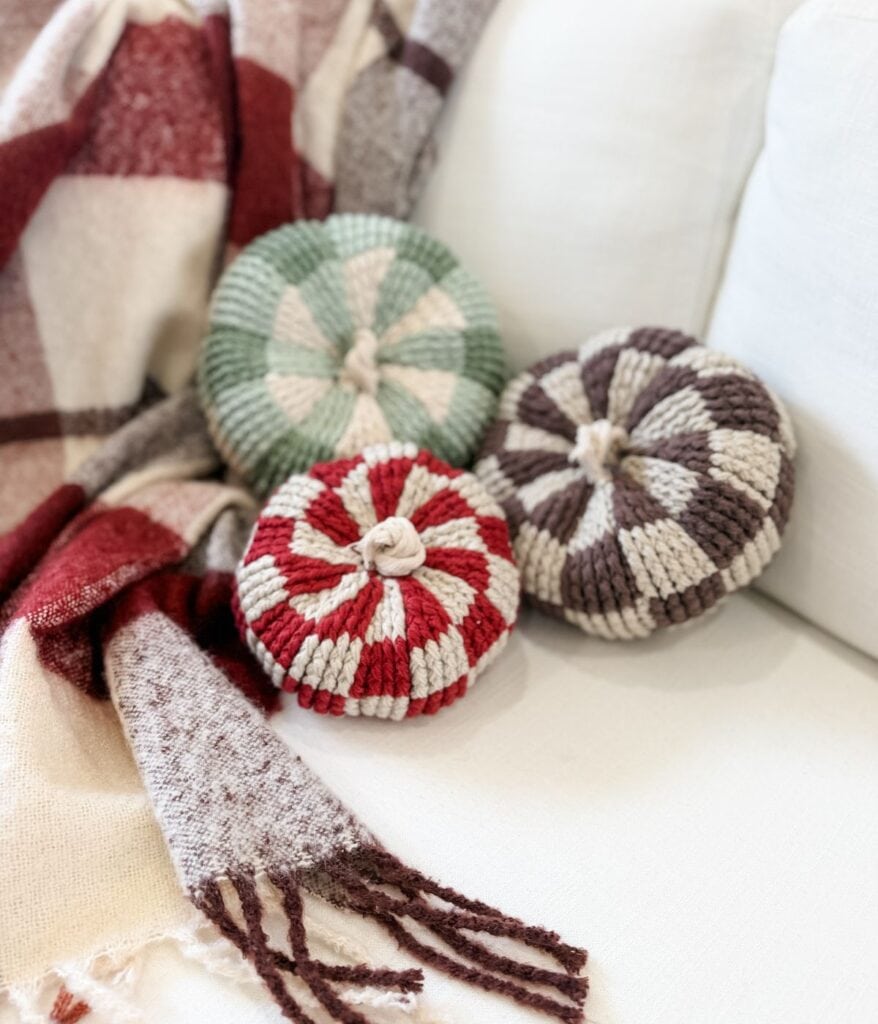 Three crocheted, pumpkin-shaped pillows in green, red, and brown gingham pumpkin patterns sit on a white sofa next to a plaid throw blanket.