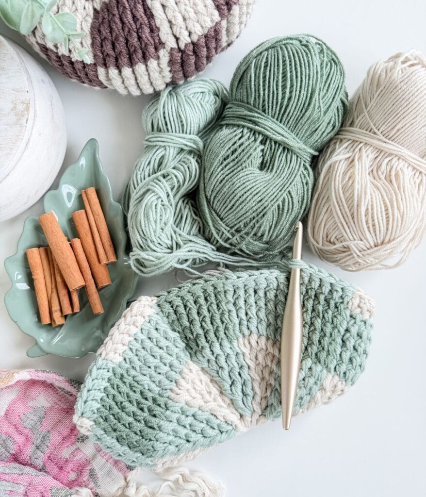 A crochet project in progress for a gingham pumpkin pattern with a gold hook, skeins of green and cream yarn, and a small dish of cinnamon sticks.