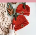 Two orange crochet pumpkin-shaped hats, each with a green stem and a leather tag, displayed next to cinnamon sticks on a white table as an inspiration for your next crochet pattern.